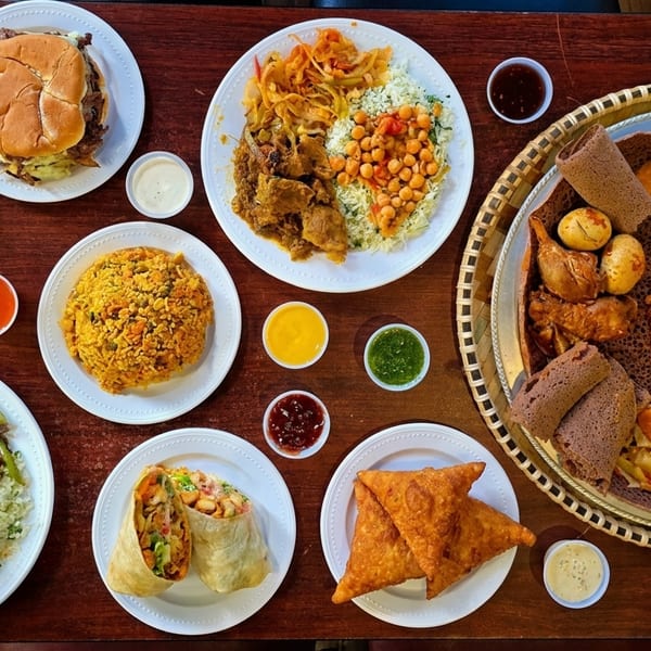 An overhead view of a wooden table laden with various Ethiopian and American dishes, including injera, rice, burgers, and samosas.